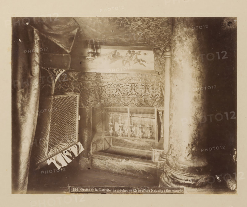 Grotte de la Nativité: la crêche, Cave of the Nativity: the manger, Cradle of Christ in the Church of the Nativity in Bethlehem, album page, photograph, This photo is part of an album., number: '893', photographer: Félix Bonfils, Bethlehem (Israel), 1867 - 1885, Palestine, paper, albumen print, height 218 mm x width 277 mm, interior of church, cradle, crib, Church of the Nativity, A dim, sepia-toned interior scene showing patterned fabric draped across the ceiling and walls, a hanging lamp suspended by chains, ornate carved columns with worn surfaces, a mesh-covered case or screen with decorative edging, a cushioned or padded surface beneath the mesh, and layered textiles with floral or damask motifs filling much of the space.
