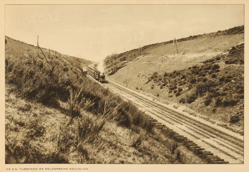 State Railways train on the tracks in the hills of Gelderland, The SS between the Gelderland hills, album page, photomechanical print, This print is part of an album., Gelderland, cliché maker: Nederlandsche Rotogravure Maatschappij, Leiden, in or before 1915, photographer, paper, rotogravure, height 161 mm x width 239 mm, railway, train, low hill country, A sepia-toned landscape showing railroad tracks running through a valley between low hills covered in grasses and shrubs, with a steam locomotive pulling cars and emitting a plume of smoke as it travels along the rails, wooden ties visible alongside the tracks, scattered telegraph poles standing on the hillsides, and dense vegetation in the foreground sloping down toward the tracks.
