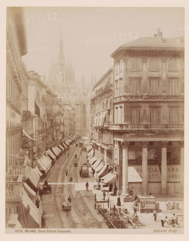 View of the Corso Vittorio Emanuele in Milan, Italy, Corso Vittorio Emanuele, Milano, photograph, number: '3830.', photographer: Giacomo Brogi, Milan, publisher: Edizioni Brogi, Florence, 1864 - 1881, Tuscany, Lombardy, pasteboard, paper, albumen print, height 330 mm x width 243 mm, urban housing, street, Corso Vittorio Emanuele II, A sepia-toned street scene showing a wide avenue flanked by tall multi-story buildings with shuttered windows and awnings, a large corner building with columns and a tiled roof on the right, tram tracks running down the center of the street, small horse-drawn carts and early motor vehicles and bicycles moving along the roadway, pedestrians walking on sidewalks and standing near storefronts, awnings and signs above ground-floor shops, scaffolding and construction activity with workers and materials visible near the bottom right, and a distant ornate cathedral façade rising above the street between the buildings.