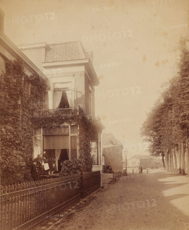 Street scene, with man, woman, and two children in front of a house (former title), Johannes Hendrikus Graadt van Roggen and his family in the doorway of their house at Lage Markt 33 in Nijmegen, photograph, Johannes Hendrikus Graadt van Roggen and his family in the doorway of their home at Lage Markt 33 in Nijmegen, annotation: 'Lange ?', stamp: 'W.Jvens (sic!), Photo aaf (?), Nijmegen', photographer: Wilhelm Ivens, Nijmegen, c. 1890 - c. 1915, paper, pasteboard, gelatin silver print, gelatin silver print on photographic paper, height 270 mm, width 220 mm, Person, Johannes Hendrikus Graadt van Roggen, A sepia-toned street scene showing the facade of a multi-story brick house partially covered in climbing vines, with tall windows and a small balcony with decorative railing and potted plants; curtains hang in the open windows and a group of people sit and stand on the ground-level porch area near a wrought-iron fence; a tree-lined street runs alongside the house with a lamp post and dis...