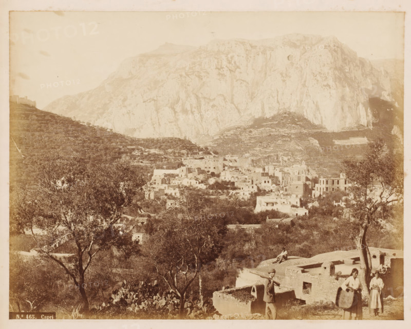 Capri, View of the city of Capri, photograph, number: 'No. 465', photographer: attributed to Roberto Rive, Isola di, 1860 - 1889, Attributed to, pasteboard, paper, albumen print, height 194 mm x width 244 mm, height 239 mm x width 277 mm, city-view in general, 'veduta', mountains, A sepia photograph of a hillside village with clustered stone and stucco buildings nestled among terraces and vegetation in front of a large rocky mountain; trees and low shrubs fill the foreground, with a few scattered cacti and a couple of figures near ruined low walls, one person standing and carrying a long pole and two women holding shallow baskets, and another figure seated on a flat roof; the mountains rise steeply behind the village with layered rock faces and light shadowing across the slopes, and a distant slope with terraced fields visible to the left.