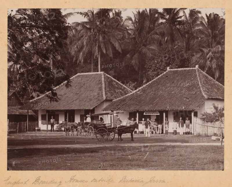 English boarding houses outside Batavia (present-day Jakarta), Java, Dutch East Indies, photograph, Dutch East Indies. Above the entrance of one of the houses hangs the sign: 'WJ Schinkel Boarding House'. In front of the houses, a couple, wagons, and people are posing. Palm trees in the background. Possibly an album page., inscription: 'English Boarding Houses outside Batavia Jarva. sic', c. 1900 - c. 1915, photographer, pasteboard, paper, gelatin silver print, height 161 mm, width 216 mm x width 289 mm, height 244 mm, West Java, Batavia, A sepia photograph shows a row of single-story buildings with tiled pitched roofs set against a backdrop of tall palm trees and dense foliage; in front of the buildings a group of people stand and sit, some wearing light-colored clothing and hats, others in darker garments, and several appear to be posed near or beside horse-drawn carriages with large wooden wheels; a wooden fence runs along one side of the scene and a dirt ground extends in the foreg...