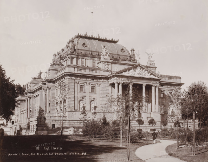 Royal Theater, Wiesbaden, Germany, photograph, View of the Royal Theater, with a park, and a man in uniform in the foreground, Germany., inscription: '112. Kgl. Theater., Mondel & Jacob, Inh. G. Jacob, Hof-Photh. Wiesbaden: 1896.', photographer: Mondel & Jacob, photographer: Mondel, photographer: Julius Jacob, 1896, Hessen, pasteboard, paper, daylight collodion silver print, height 226 mm, width 281 mm x width 330 mm, height 510 mm, A large ornate stone building with a domed roof and decorative sculptures along its roofline and pediments occupies the center of the image, featuring tall columns at its entrance, carved reliefs above the columns, and sculpted figures on ledges and corners; trees and trimmed shrubs stand in front of the building along a curved pathway where a person in a dark coat and light trousers walks, and lamp posts and a stone statue on a pedestal appear near the steps leading up to the building.