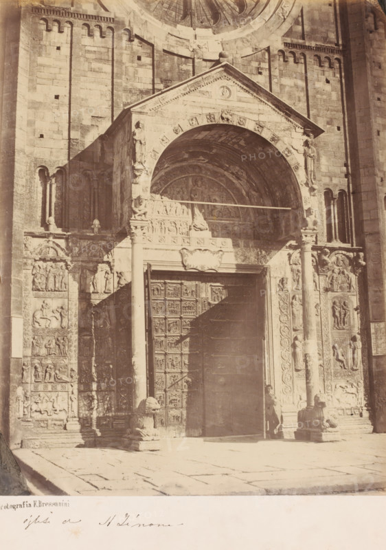 Portal of the Basilica di San Zeno (also: San Zeno Maggiore), Verona, Italy, photograph, Italy., date: '1860's', stamp: 'Fotografia E. Bressanini', caption: 'ogliti di St. Zinone ?', number: '1183.', photographer: Emilio Bressanini, c. 1865 - c. 1880, Emilio Bressanini, photographer, pasteboard, paper, albumen print, height 276 mm x width 218 mm, height 411 mm x width 301 mm, S. Zeno, A sepia-toned photograph of an ornate stone entrance with a large arched portal set into a brick façade, flanked by carved columns and sculpted relief panels; the arch contains decorative carvings and a recessed semicircular tympanum with figures, and a wide painted shadow crosses the doorway and surrounding stonework; tall carved doors with square panels occupy the central opening, and sculpted lion figures sit at the base of the columns on either side of the steps; a person stands near the right column partially in shadow; the ground in front is paved with large stone slabs and the upper wall shows rows...
