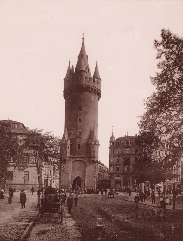 Eschenheimner Turm, Frankfurt-am-Main, Germany, photograph, Germany., caption: 'Frankfurt', c. 1875 - c. 1900, photographer, paper, pasteboard, collotype, photoprint on photo paper on cardboard, height 274 mm, width 208 mm x width 490 mm, height 330 mm, Hessen, Frankfurt am Main, A tall cylindrical tower with conical turrets and a large arched gateway stands at the center of a street scene, flanked by multi-story buildings with ornate facades and a cluster of trees with leafy branches on the right; cobblestone streets fill the foreground with tram tracks running down the middle, horse-drawn carriages parked near the curb, and numerous people walking or standing in period clothing scattered across the scene.