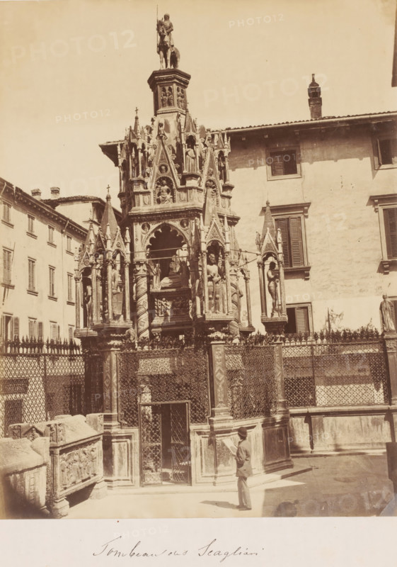 Tomb of the Scagliari family, Italy, photograph, Tomb in the city, with an ornately decorated fence and a man, tourist? with a bowler hat in front of it, reading information about the monument ?., caption: 'Tombeau ons ? Scagliari', number: '1182', inscription: '1860's BRESSANINI', c. 1865 - c. 1880, photographer, pasteboard, paper, albumen print, albumendruk, height 280 mm x width 218 mm, height 415 mm x width 300 mm, A tall ornate stone monument with multiple pointed arches, sculpted figures, and decorative columns rises from a fenced base in a courtyard, surrounded by buildings with shuttered windows; the monument features a mounted figure at its highest point, niches with statues, carved reliefs, and intricate metalwork on the surrounding railing, while a person in a suit and hat stands near the base looking up.