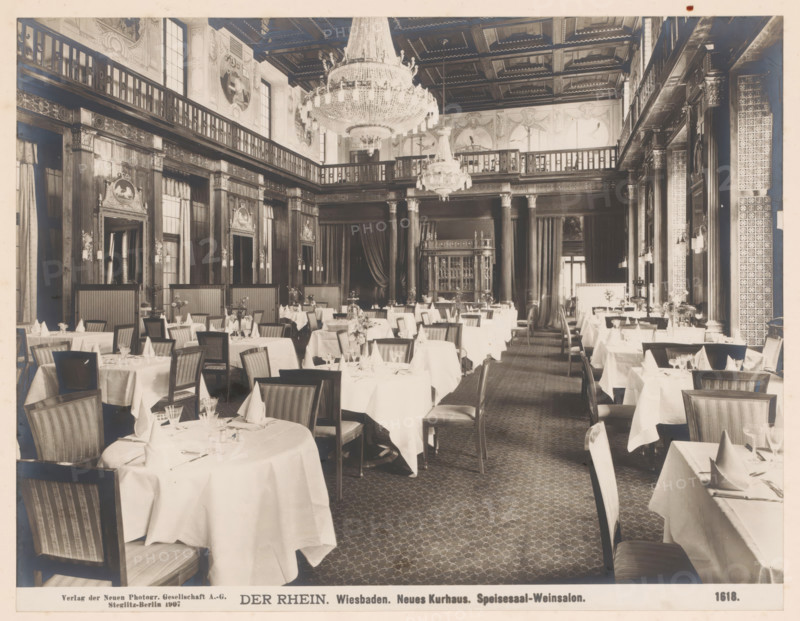Speisesaal, Weinsalon, Neues Kurhaus, Wiesbaden, Germany, photograph, Germany., caption: 'DER RHEIN. Wiesbaden. Neues Kurhaus. Speisesaal-Weinsalon.', name: 'Neue Photographische Gesellschaft AG, Steglitz-Berlin 1907', number: '1618.', photographer: Neue Photographische Gesellschaft, Berlin, 1907, pasteboard, paper, albumen print, albumendruk, height 189 mm, width 245 mm x width 328 mm, height 510 mm, Hesse, A wide dining room with cloth-covered round and rectangular tables arranged in rows, each table set with folded napkins, glassware, and small floral centerpieces, surrounded by wooden chairs with upholstered seats and striped backs; tall decorative columns and wood-paneled walls rise to a balcony level with balustrades, ornate carved moldings, framed medallion reliefs, and draped curtains; two large crystal chandeliers hang from a coffered ceiling, and patterned carpeting covers the floor.