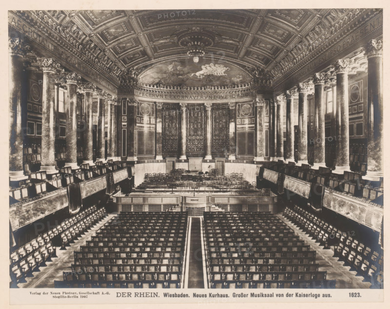 Der Rhein. Neues Kurhaus, Grosser Musiksaal von der Kaiserloge aus, Wiesbaden, Germany, photograph, Germany., caption: 'DER RHEIN. Wiesbaden. Neues Kurhaus. Grosser Musiksaal von der Kaiserloge aus.', name: 'Neue Photographische Gesellschaft AG, Steglitz-Berlin 1907', number: '1623.', photographer: Neue Photographische Gesellschaft, Berlin, 1907, pasteboard, paper, albumen print, albumendruk, height 192 mm, width 239 mm x width 328 mm, height 510 mm, Hesse, A large ornate concert hall with rows of seating arranged facing a raised stage area, flanked by tall decorative columns with carved capitals and ornate paneling along the walls, a curved apse behind the stage featuring intricate relief panels and decorative doors, an elaborately coffered ceiling with sculpted moldings and a central chandelier, balcony seating with carved balustrades on both sides, and many wooden chairs arranged on the stage and in the auditorium.
