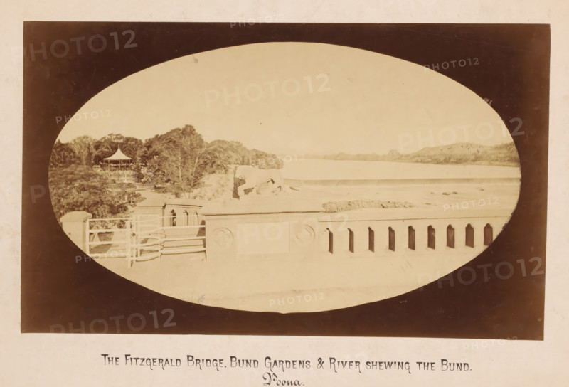 Fitgerald Bridge, Bund Gardens and Mula Mutha river, Pune, Maharashtra, India, photograph, Oval photo of a bridge with river and a park on the left. Poona, also called Pune and Puna is an Indian city not far from Mumbai., inscription: 'The Fitzgerald Bridge + Bund Gardens + River shweing the Bund, Poona', inscription: 'THE FITZGERALD BRIDGE, BUND GARDENS & RIVER SHEWING THE BUND., Poona.', photographer: X photo., c. 1875 - c. 1900, An oval sepia photograph framed by a dark rectangular border shows a riverside scene with a low balustrade in the foreground featuring decorative cutouts and carved circular motifs, a set of railings and a small stair or platform area, a row of trees and dense foliage to the left with a pavilion topped by a pointed roof visible among the trees, a low embankment leading to a wide calm river that stretches toward distant low hills under a pale sky, and faint signs of wear and spotting on the photographic surface.