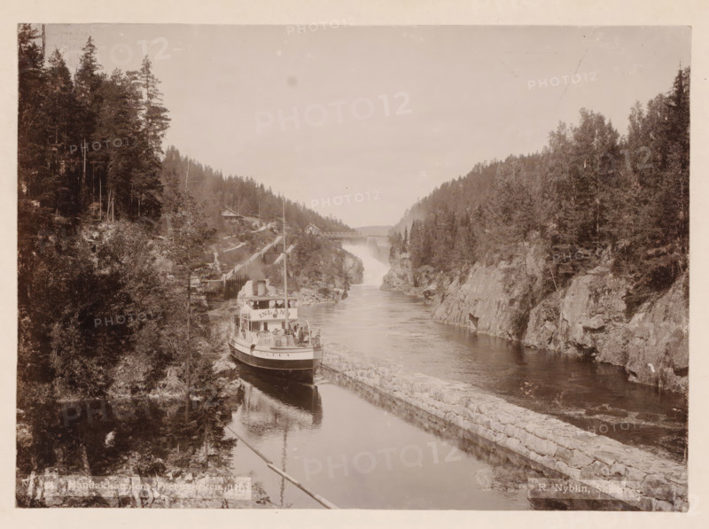 Vrangfos, Telemarken, photograph, Frontal view of a boat passing through a lock, with the river alongside. The Telemark Canal (Telemarkskanalen) in Norway consists of two canals, of which the Norsjø-Skien Canal with locks in Skien and Løveid, built between 1854 and 1861, is the oldest of the two canals. The other canal we see in the photo is the Bandak-Norsjø Canal, opened in 1892 =terminus post quem., caption: 'No. 5 Vrangfos, Telemarken sic., R. Nyblin, Skien., Eneberettiget 1896.', photographer: Ragnvald Nyblin, Skien, in or after 1896 - c. 1915, Telemark, paper, pasteboard, daylight collodion silver print, daylight collodion silver print on photo paper on cardboard, height 157 mm, width 218 mm x width 311 mm, height 380 mm, A sepia-toned scene of a narrow river flanked by steep rocky banks covered in dense trees, with a passenger steamship moored along a stone-lined channel near the left bank, its upper deck populated by people and railings and a tall smokestack rising above; a win...