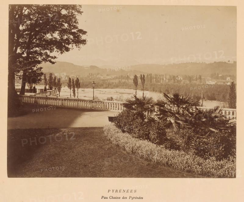 Pyrénées, Peau Chaine des Pyrénées', Pyrenees, France, photograph, View from a park over the landscape, with houses and mountains in the background., date: '8, 10 October 1884.', inscription: 'Pyrénées, inscription: 'Papeterie & Librairie, LAFON, 3. Rue Henry IV. 3, PAU, EAUX-BONNES, Etablissement Thermal', vendor: Lafon, Pau, 1884-10-08 - 1884-10-10, photographer, pasteboard, paper, albumen print, albumendruk, height 192 mm, width 253 mm x width 385 mm, height 340 mm, A sepia-toned scene showing a riverside landscape viewed from a terrace with a balustrade, framed at left by large tree trunks and leafy branches and at right by palms and dense shrubs, with a paved area and manicured lawn in the foreground, a lamp post near the railing, slender trees along the riverbank, a wide river beyond them, rolling hills with scattered buildings and tree lines in the middle distance, and a pale sky above.