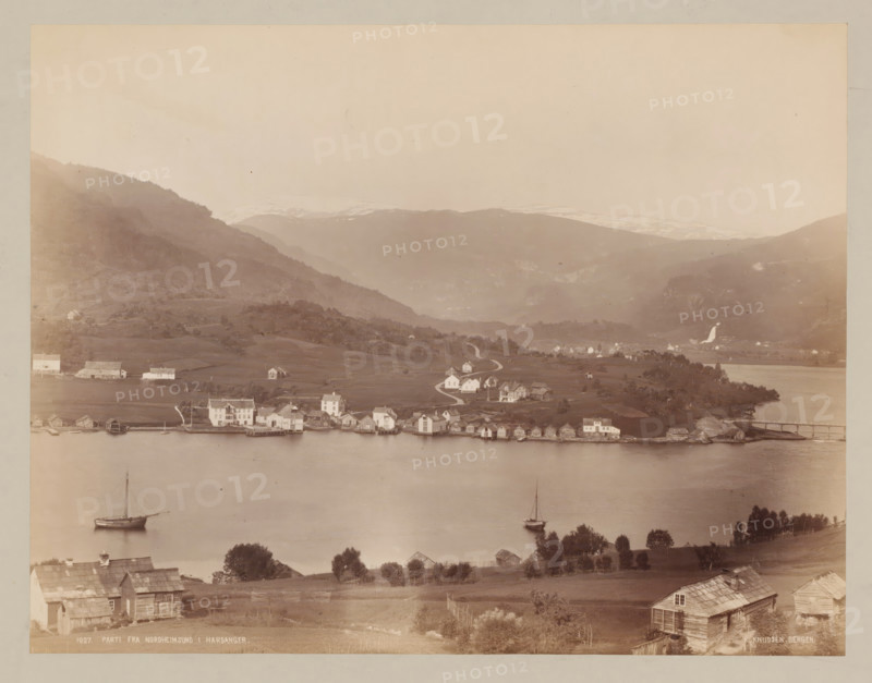 Parti fra Nordheimsund, Hardangerfjord ?, Norway, photograph, Norway., inscription: 'K. KNUDSEN, BERGEN', caption: '1027. PARTI FRA NORDHEIMSUND I HARDANGER.', photographer: Knud Knudsen, c. 1880 - c. 1900, paper, pasteboard, albumen print, height 209 mm, width 272 mm x width 402 mm, height 316 mm, A panoramic view shows a calm body of water in the foreground with two sailboats anchored near the shore, a shoreline lined with small wooden boathouses and larger white buildings, gently rolling grassy land with scattered trees and fences rising behind them, a winding road leading up to more clustered houses spread across the meadow, steep wooded hills and terraced slopes rising further back, distant mountains with pale snow patches near their peaks, and a bridge connecting a small promontory to the far shore.