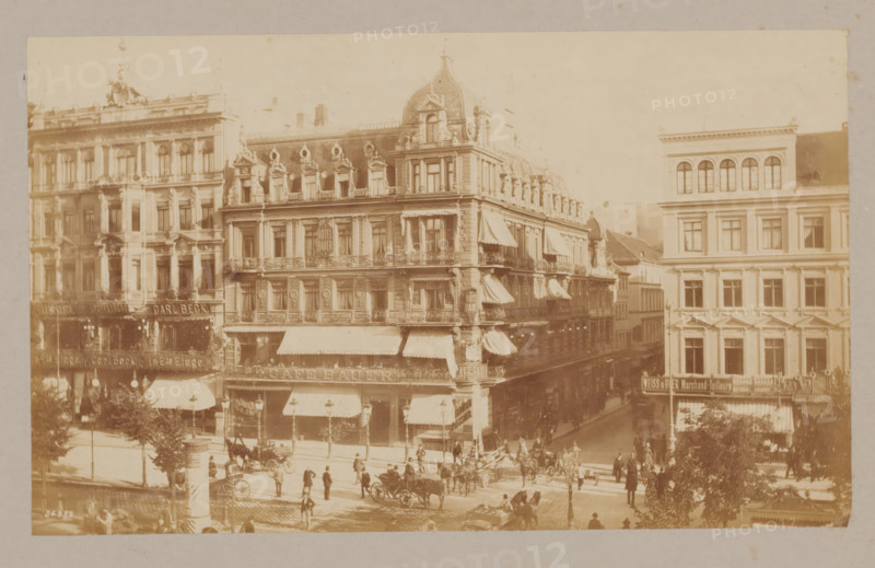Café Bauer, Unter den Linden, Berlin, photograph, Berlin., inscription: 'Berlin', number: '24989', c. 1880 - c. 1900, photographer, paper, pasteboard, albumen print, albumendruk, height 158 mm, width 261 mm x width 406 mm, height 308 mm, A wide sepia-toned street scene shows several multi-story buildings with ornate facades, decorative windows, balconies, and awnings, with one prominent corner building featuring a domed roof and multiple small dormer windows along its upper level; horse-drawn carriages and carts are visible on the cobblestone street below, along with groups of people walking or standing on the sidewalks and near the intersection, street lamps and trees lining the thoroughfare, and architectural details such as columns, sculpted ornamentation, and ironwork railings on the buildings.