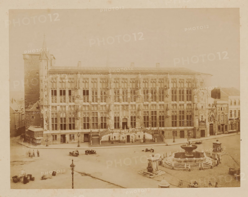 Aachen City Hall after the fire', Germany, photograph, 'Aachen City Hall after the fire', Germany., number: '19 (in the same ink, handwriting as inscription verso) with number: 38 written in pencil over it', inscription: 'Stadhuis v. Aken na den brand', in or after 1883 - c. 1910, photographer, pasteboard, paper, albumen print, albumendruk, height 180 mm, width 230 mm x width 381 mm, height 304 mm, North Rhine-Westphalia, Aachen, A wide sepia-toned photograph of a large ornate stone building with many tall rectangular windows and decorative stonework across its façade, a broad central staircase with balustrades leading to an entrance, lamp posts along the street in front, a multi-tiered circular fountain with water basins and surrounding railings in the square, a tall tower attached to the left side of the building and smaller adjoining structures on the right, several small groups of people and a few horse-drawn vehicles scattered on the open paved area, and various architectural deta...