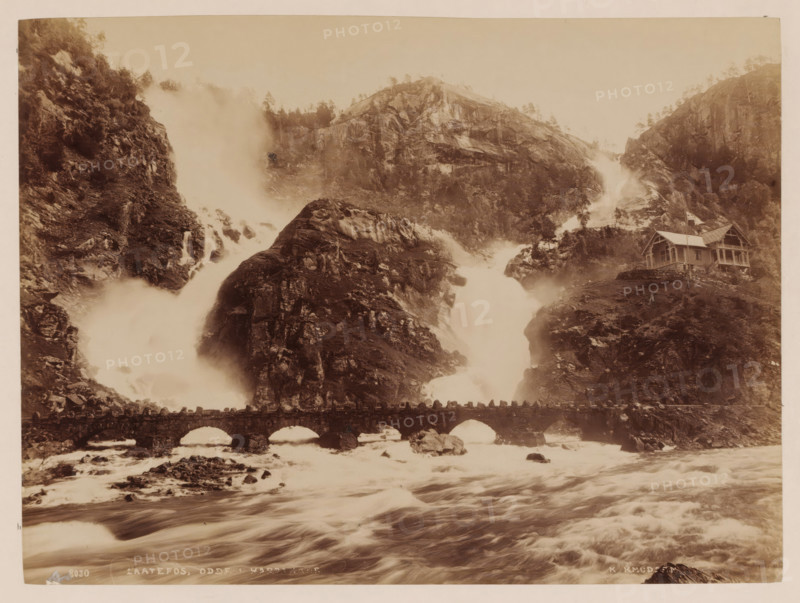 Laatefos, Hardanger, Norway, photograph, Two wild rivers flow from a mountain together into a large river in the foreground, Norway., inscription: 'K. KNUDSEN ...', caption: '8030. LAATEFOS. ODDF ? HARDANGER', photographer: Knud Knudsen, c. 1880 - c. 1900, paper, pasteboard, albumen print, albumendruk, height 163 mm, width 218 mm x width 311 mm, height 396 mm, A wide sepia-toned landscape shows steep rocky cliffs with water cascading down in powerful streams and mist rising where the falls meet the river below, a stone arched bridge crossing the turbulent water in the foreground, and a wooden house with a pitched roof perched on a rocky slope to the right, surrounded by scattered trees and rugged terrain.