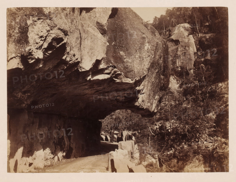 Parti ved halvtunnellen I Bratlandsdalen, Suldal, Norway, photograph, Tunnel of overhanging rocks., inscription: 'K. Knudsen, Bergen.', inscription: '8380. Parti ved halvtunnellen I Bratlandsdalen, Suldal', photographer: Knud Knudsen, Bergen, c. 1880 - c. 1900, paper, pasteboard, albumen print, height 160 mm, width 217 mm x width 310 mm, height 402 mm, A sepia-toned photograph showing a rocky cliff face with a dark overhanging ledge above a narrow dirt road, jagged rock formations lining the road and a low stone barrier along the roadside, a few standing figures on the road near the stone barrier, a steep wooded slope with trees and shrubs rising beside the cliff, and textured rock surfaces with light and shadow across the scene.