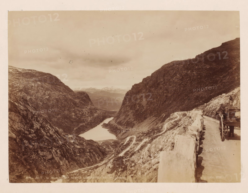 View from Reldalsfjeldet with mountains around it, Norway, photograph, View over a deep valley surrounded by mountains, taken from a road with a cart next to it. Photographer appears to have dismounted., inscription: 'View from Reldalsfjeldet with mountains around it', inscription: 'K. Knudsen, Bergen.', photographer: Knud Knudsen, Bergen, c. 1880 - c. 1900, paper, pasteboard, albumen print, height 163 mm, width 218 mm x width 310 mm, height 386 mm, A sepia landscape photograph showing a narrow valley between steep rocky cliffs with a winding road carved along the cliffside and a river or lake running through the valley floor, a stone barrier lining the road in the foreground, a vehicle partially visible on the road at the right edge, and distant mountains and cloudy sky in the background.