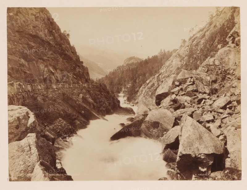 Parti Af Oddedalen, Norway, photograph, View of a wild flowing river between mountains and rocks, Norway., inscription: 'Udisgt fra Reldalsfjeldet nedover gaarsbottnen mod folgffonden', inscription: 'K. Knudsen, Bergen.', photographer: Knud Knudsen, Bergen, c. 1880 - c. 1900, paper, pasteboard, albumen print, albumendruk, height 163 mm, width 216 mm x width 316 mm, height 400 mm, A sepia-toned landscape showing a rocky river gorge with steep cliff faces on both sides and large boulders scattered along the riverbank, water flowing through the narrow channel appearing smooth and misty, coniferous trees clinging to the slopes and ridge lines, a rough stone retaining wall or path built into the cliff on the left side, distant hazy hills fading into the sky, and a foreground of angular rocks and ledges descending toward the water.