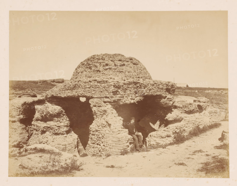 Canopy, house of wood stones?, with a man in front of it, photograph, with a man in front of it., inscription: 'Ph. J Carrianes ?', c. 1870 - c. 1900, photographer, pasteboard, paper, albumen print, height 197 mm, width 262 mm x width 443 mm, height 325 mm, A sepia photograph showing a large, rounded rock formation with layered, weathered textures and several deep crevices; the rock occupies the central foreground and middle distance, with smaller rocky outcrops and patches of sparse vegetation around its base and extending into the background; a man wearing a dark jacket, light trousers, and a hat is seated in a recess of the rock near the lower center of the image; the horizon and flat landscape stretch behind the rock under a clear, pale sky.