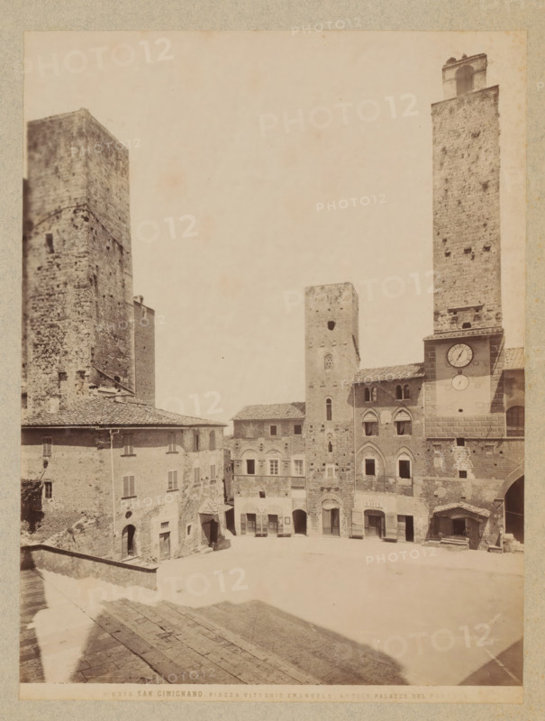 Piazza Vittorio Emanuele, Antico Palazzo del Podesta', San Gimignano, Tuscany, photograph, The Antico Palazzo del Podestà and the Torre della Rognosa tower in San Gimignano, located in front of the Basilica., inscription: '6376 San Gimignano - Piazza Vittorio Emanuele - Antico Palazzo Del Podesta', inscription: 'NORWAY', c. 1870 - c. 1900, photographer, pasteboard, paper, albumen print, height 190 mm x width 250 mm, height 444 mm x width 356 mm, A pale sepia photograph of a town square bordered by tall stone towers and multi-story masonry buildings with tiled roofs, showing textured brick and stone facades, arched windows and doorways, shuttered windows, a large clock set into one tower, and a small awning over a shop entrance; the foreground shows worn stone steps and a sloped paved surface leading down into the open square, while shadows fall across the pavement and parts of the buildings.