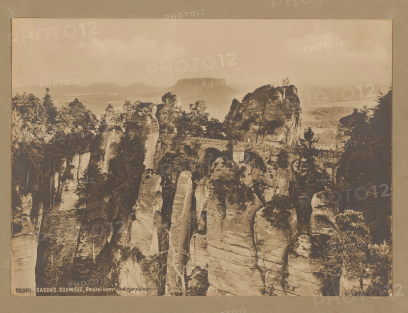 Bastei from the Ferdinandstein, Saxon Switzerland, Germany, photograph, Saxon Switzerland (Saxon Switzerland) is a former district (Landkreis) in the German state of Saxony and also the name for the region in the Elbe Sandstone Mountains between Dresden and the Czech border. Here we see the 'Bridge of Bastei', a natural wonder of several striking rock peaks with a man-made stone bridge., number: '1903', inscription: '10, 001 SAECHS. SCHWEIZ, Bastei vom Ferdnindstein.', check stamp: 'Th. ...-maier', 1903, photographer, pasteboard, paper, gelatin silver print, developing gelatin silver print, height 169 mm, width 230 mm x width 381 mm, height 320 mm, A sepia-toned photograph showing a rocky landscape with tall vertical sandstone pillars and cliffs, many topped with trees and vegetation, and a stone bridge spanning between rock formations; the foreground displays steep weathered rock faces with visible striations and sparse trees growing from crevices, while the midground features cluster...