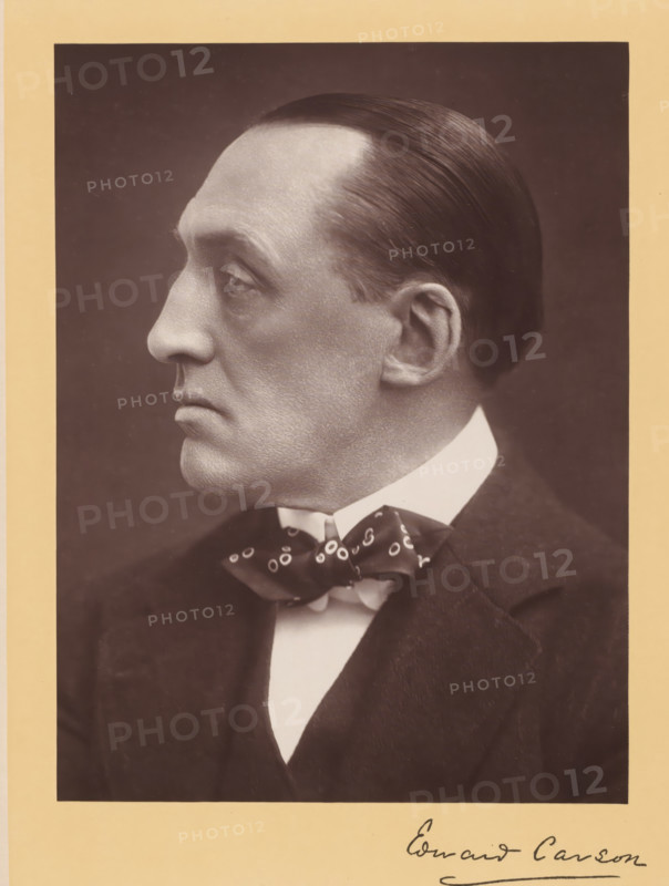 Sir Edward Henry Carson, photograph, Portrait of a man and profile, caption: 'The Right Honorable Sir Edward Henry Carson, KC, LL.D.', caption: 'Russell & Sons, Photographers, Copyright., 17, Baker St., London W.', caption: 'Edward Carson', photographer: Russell & Sons, London, c. 1890 - c. 1910, pasteboard, paper, carbon print, height 225 mm, width 170 mm x width 263 mm, height 366 mm, A sepia-toned portrait of a man in profile facing left, showing a smooth, slicked-back hairstyle, a pronounced nose, a high forehead, and a clean-shaven face with visible skin texture; he is wearing a white shirt with a high collar, a patterned bow tie, and a dark suit jacket, and the photograph is mounted on a pale mat with part of a handwritten signature visible at the bottom edge.
