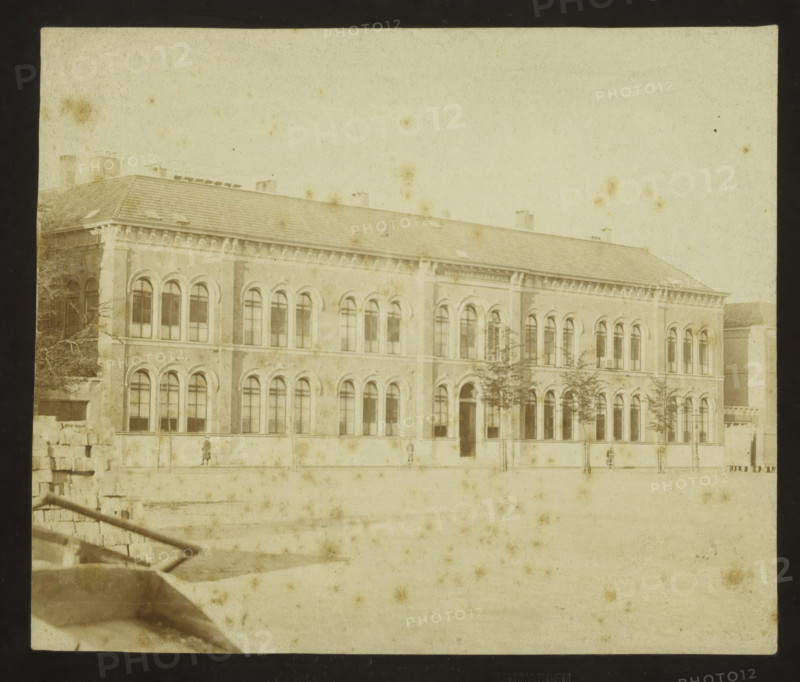 View of the former secondary school in Leeuwarden, photograph, inscription: 'HJ Craije Azn. LEEUWARDEN', photographer: Hendrik Johannes Craije Azn., Netherlands, 1879 - 1910, paper, pasteboard, albumen print, height 122 mm x width 145 mm, height 140 mm x width 190 mm, façade (of house or building), Leeuwarden, A faded, sepia-toned photograph of a large two-story brick building with a sloped tiled roof and multiple chimneys, featuring rows of tall arched windows on both levels and decorative cornices along the roofline, with a few small trees planted in front of the building and a mostly empty paved area in the foreground, a low stone wall and steps visible on the left side, and a couple of small human figures standing near the facade.