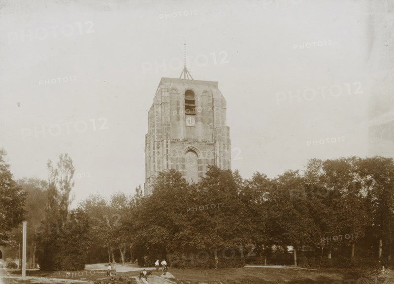 View of the Oldehove with nearby park in Leeuwarden, photograph, Leeuwarden, 1890 - 1910, photographer, paper, pasteboard, daylight collodion silver print, height 123 mm x width 170 mm, height 144 mm x width 194 mm, parts of church exterior and annexes: tower, A tall stone tower with arched openings and a pointed finial rises above a dense line of trees, the tower showing vertical brick patterns and a large arched recess near its base; in front of the trees a grassy area contains a few people standing and sitting along a low stone edge and a small rectangular water feature, while tall slender trees flank the scene and the sky above is pale and empty.