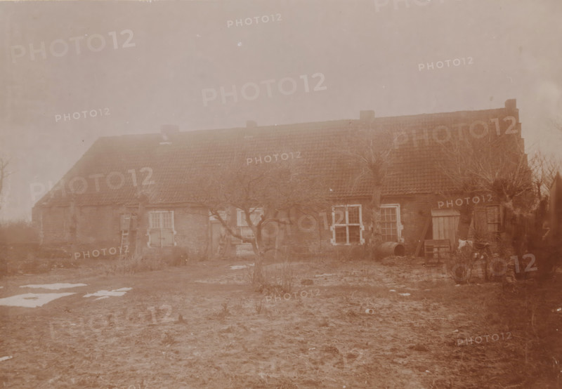 Aytta State (house of God or church) in Swichum, photograph, annotation: 'Aytta State House in Swichem, Ph ? Dec 1909, Friesland', Swichum, photographer: AJM Mulder (possibly), 1909, Attributed to, paper, pasteboard, daylight collodion silver print, height 150 mm x width 211 mm, church (exterior), A long, low brick building with a tiled roof and several chimneys stretches across the scene, fronted by leafless, gnarled trees and bare ground with patches of snow; there are multiple windows with visible panes, a few wooden doors, a ladder leaning against the wall, a barrel and scattered wooden boards near one end, and some low vegetation and rough earth in the foreground.