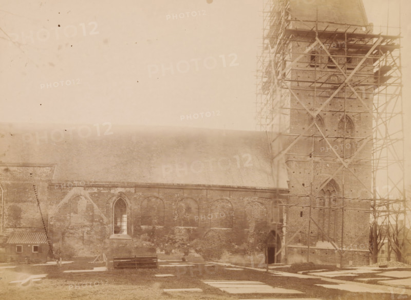 North side of the St. Vitus Church in Stiens, during restoration work, photograph, annotation: 'Ned. Herv Kerk te Stiens fr (Fr.), North side, Nov 1898', annotation: 'Leeuwarderadeel, Stiens', Stiens, photographer: AJM Mulder (possibly), 1898, Attributed to, paper, pasteboard, albumen print, height 164 mm x width 225 mm, church (exterior), Sint-Vituskerk (Stiens), A sepia-toned photograph showing a large brick building with arched windows and a steep roof, a tall tower at one end surrounded by wooden scaffolding, a small tiled-roof outbuilding near the corner, visible grave markers or flat stones on the grassy foreground, a wooden bench and some construction materials near the base of the building, and a faintly mottled sky above.