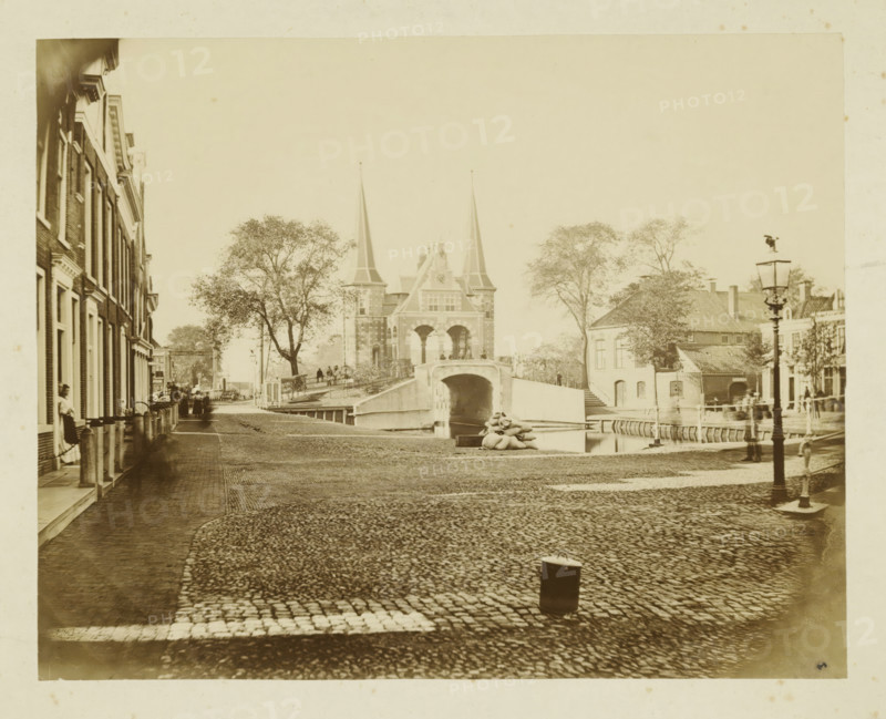 View of Sneek with the city side of the Sneeker Waterpoort, photograph, annotation: 'f1, - ..., Sneek', Sneek, 1870 - 1900, photographer, paper, pasteboard, albumen print, albumendruk, height 145 mm x width 182 mm, height 255 mm x width 286 mm, city-gate (city(-scape) with figures, staffage), Waterpoort (Sneek), A wide cobblestone street slopes toward a stone arch bridge flanked by two tall pointed towers, with trees on either side and people visible on the bridge and along the banks, a canal running beneath the bridge where a small boat is moored, brick row houses lining the left side of the street with several figures standing near doorways, a lamppost in the foreground on the right, low buildings and more trees beyond the bridge, and a pile of sacks near the canal edge.