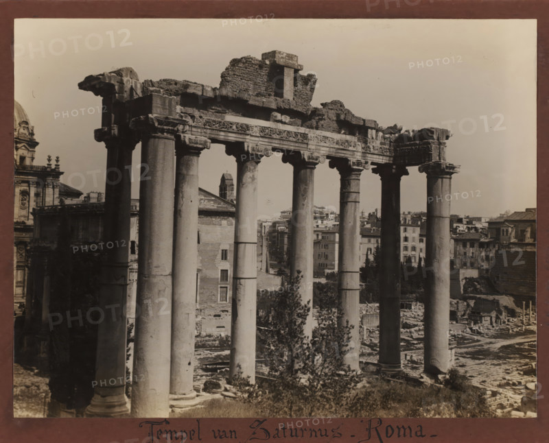 Temple of Saturn, Rome, Roman Forum, Italy, album page, photograph, Italy., inscription: 'Temple of Saturn - Roma', c. 1900 - c. 1940, photographer, pasteboard, paper, albumen print, height 185 mm x width 242 mm, height 207 mm x width 263 mm, A sepia-toned photograph of a row of tall stone columns with ornate capitals supporting a partially ruined entablature, with broken masonry and rubble atop the structure; behind and between the columns are glimpses of older buildings, rooftops, and a bell tower rising above surrounding structures; the foreground shows uneven ground, scattered stones, and vegetation at the base of the columns; to the left a portion of a domed building with decorative architectural details is visible, and to the right more urban buildings extend toward the horizon under a clear sky.