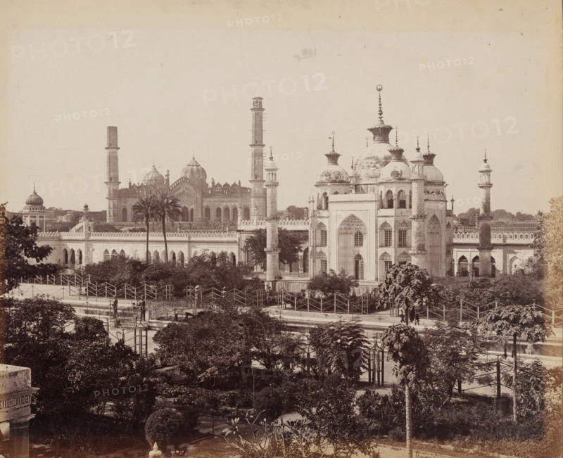 View of mosques in Lucknow, Uttar Pradesh, India, photograph, inscription: 'Lucknow', number: '35', signature: 'Bourne 1051', photographer: Samuel Bourne, c. 1863 - c. 1868, paper, albumen print, height 234 mm x width 284 mm, Oudh, Lucknow, A wide sepia photograph of an ornate building complex with multiple domes and tall slender towers rising above a long crenellated wall, palm trees and dense foliage in the midground and foreground, arched windows and decorative entrances on the nearer structures, domes topped with finials and bulbous crowns, latticework and balustrades along the rooftop edges, paved walkways and fenced pathways weaving through landscaped gardens, lampposts and a few small human figures near the pathways, and a clear sky occupying the upper portion of the image.