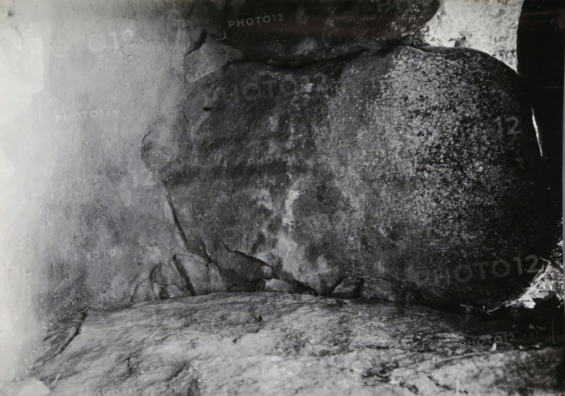 Keystone of the Kerguntuil dolmen, Trégastel, photograph, inscription: 'right - the keystone of the original, dolmen., Allée couverte de Kergantuil, Trégastel. C. du N.', photographer: attributed to Jan Lanting, supervision: Albert Egges van Giffen, 1939-05-27 - 1939-06-04, Attributed to, paper, baryta paper, gelatin silver print, developing gelatin silver print, height 123 mm x width 170 mm, megalithic chambered tomb, dolmen, A close-up view of a rough stone surface filling the frame, showing textured rock planes with cracks, crevices, and layered formations; areas of lighter and darker shading create mottled patterns, with one side appearing speckled and another side smoother; small fragments and chips of rock are visible along edges, and a narrow sliver of bright light or reflection appears near one edge; overall the image shows natural stone surfaces with varied textures and tones.