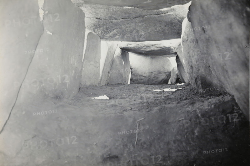 Kerguntuil Dolmen, Trégastel, photograph, inscription: 'afdr, Ban v He, Het interieur van de, allée couverte de Kergantuil, Trégastel C. du N., ZO richting', photographer: attributed to Jan Lanting, supervision: Albert Egges van Giffen, 1939-05-27 - 1939-06-04, Attributed to, paper, baryta paper, gelatin silver print, developing gelatin silver print, height 119 mm x width 170 mm, megalithic chambered tomb, dolmen, A low-angle, grayscale view of a narrow stone passage formed by large flat slabs serving as floor, side walls, and a horizontal capstone, with rough textured surfaces, scattered lighter patches on the ground, and bright light visible through the far end of the passage.