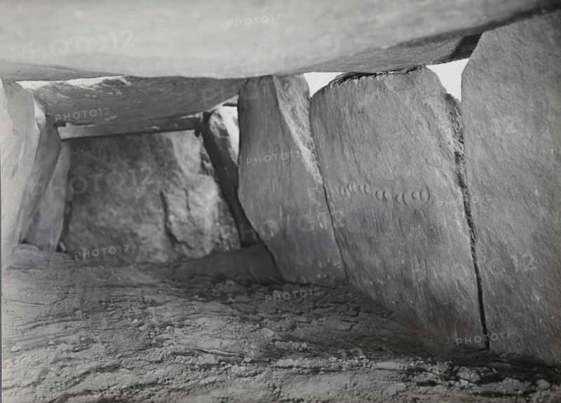 Kerguntuil Dolmen, Trégastel, photograph, inscription: 'Allée couverte de Kerganteul, Trégastel C. du N., carrying stone with the necklace (?), NW direction', photographer: attributed to Jan Lanting, supervision: Albert Egges van Giffen, 1939-05-27 - 1939-06-04, Attributed to, paper, baryta paper, gelatin silver print, developing gelatin silver print, height 123 mm x width 166 mm, megalithic chambered tomb, dolmen, A low, narrow space formed by large rough stone slabs with a textured dirt floor, the slabs showing visible striations and irregular surfaces, one vertical slab on the right featuring a row of shallow crescent indentations, light entering from a small gap near the top center and casting soft shadows along the interior, the ceiling slab appearing smooth compared to the sides, and scattered small debris and uneven patches across the ground.