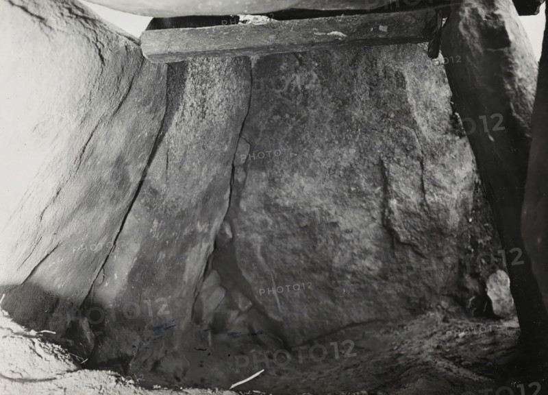 Kerguntuil Dolmen, Trégastel, photograph, inscription: 'the keystone seen from the inside, on the left the original corner filling, Allé couverte de Kerganteul, Tregastel. C. du Nord, afd, Ban dH...', photographer: attributed to Jan Lanting, supervision: Albert Egges van Giffen, 1939-05-27 - 1939-06-04, Attributed to, paper, baryta paper, gelatin silver print, developing gelatin silver print, height 122 mm x width 168 mm, megalithic chambered tomb, dolmen, A close view of rough stone surfaces forming an enclosed space, with large flat slabs leaning and meeting at angles, visible texture and cracks in the rock, a horizontal wooden beam resting near the top edge, small rounded stones tucked between slabs, and scattered dark and light patches across the uneven floor and walls.