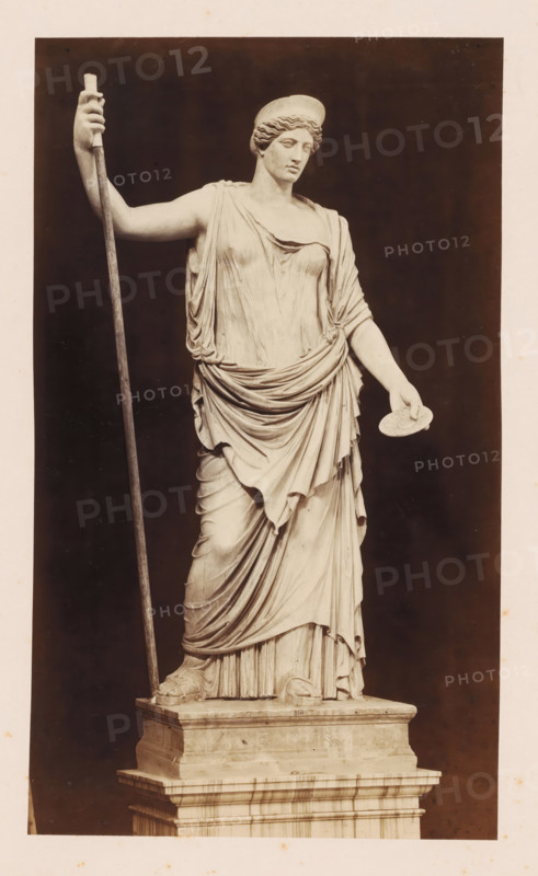 Sculpture of Juno, Museo Vaticano, photograph, photographer: Libreria Spithöver, c. 1850 - c. 1870, pasteboard, paper, albumen print, height 329 mm x width 216 mm, piece of sculpture, reproduction of a piece of sculpture, Vatican City, Vatican Museums, A full-length marble statue of a draped female figure standing on a stepped rectangular plinth is shown against a dark background, wearing a flowing, layered gown with detailed folds and a belted drapery at the waist, her left arm extended slightly forward holding a round, flat object, her right arm raised and grasping a long staff that reaches from the plinth to above her shoulder, her head turned slightly to one side and topped with a rounded headpiece, and her feet visible with sandals resting on the base.