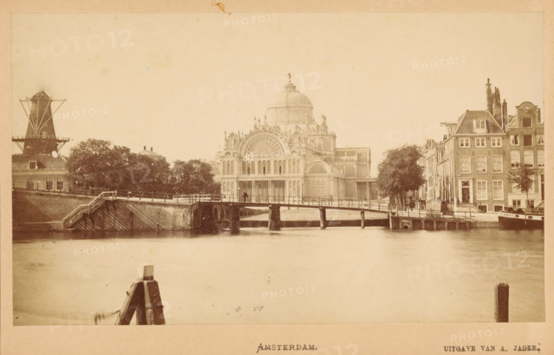 Amsterdam, View of the Palace of Popular Industry in Amsterdam, photograph, publisher: Andries Jager, c. 1865 - c. 1880, photographer, paper, pasteboard, albumen print, height 90 mm x width 145 mm, temporary exhibition, 'Salon', canal, façade (of house or building), A sepia-toned riverside scene showing a wide stretch of water in the foreground, a wooden post projecting from the water near the bottom edge, and a low arched bridge crossing the river toward the middle ground. Behind the bridge a large ornate building with a central dome and detailed arched windows dominates the center, flanked by trees on the left and a row of narrow multistory buildings with steep roofs on the right. A windmill structure is visible at the far left, and small boats and dock structures line the right bank near the row of buildings. The sky appears pale and empty above the scene.