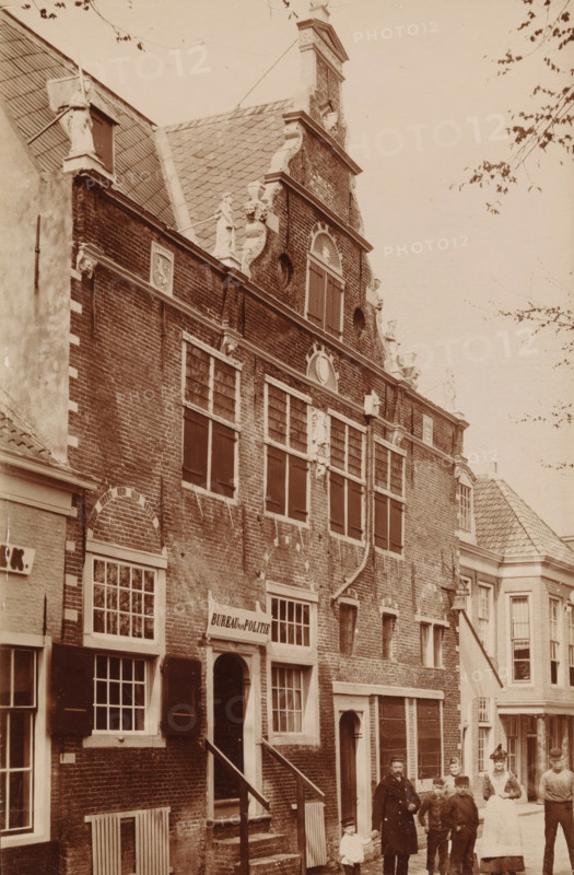 Facade of the Weigh House in Enkhuizen, photograph, inscription: 'Enkhuizen', Enkhuizen, c. 1885 - c. 1910, photographer, paper, pasteboard, daylight gelatin silver print, height 147 mm x width 97 mm, weigh-house, façade (of house or building), Waag (Enkhuizen), A brick building with stepped gables and decorative sculpted figures along the roofline fills most of the image, showing multiple shuttered windows and rectangular panes, arched brickwork above some windows, a tiled roof with a dormer, and ornamental stonework on the facade; a small flight of stairs leads to an arched doorway with a sign above it, and adjacent buildings with tiled roofs and sash windows are visible; a group of people stands on the street in front of the building, including an adult man holding a small child's hand, other children, and a woman wearing a long skirt and blouse, with branches and sparse leaves of a tree visible at the top and right edges of the scene.