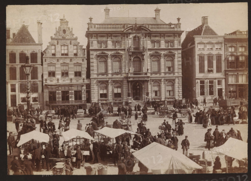 Buildings along the Grote Markt in Groningen, photograph, inscription: 'Groningen', name: 'JG Kramer Groningen', photographer: Johannes Gerardus Kramer, Grote Markt (Groningen), c. 1880 - c. 1900, Groningen (city), paper, pasteboard, albumen print, height 96 mm x width 139 mm, square, circus, façade (of house or building), A wide street scene shows a row of multi-story stone buildings with ornate facades and large windows, the central building featuring arched windows, decorative balconies, and a grand entrance with steps. In front of the buildings a busy open market fills the foreground with numerous canvas tents and stalls, carts, wooden crates, and barrels. Many people in period clothing stand, walk, or gather in small groups across the square, some near the stalls and others by horse-drawn carts. A tall, decorative lamppost stands near the left side of the image, and a few bicycles and handcarts are visible among the crowd. The overall tone of the photograph is sepia, with visible ...
