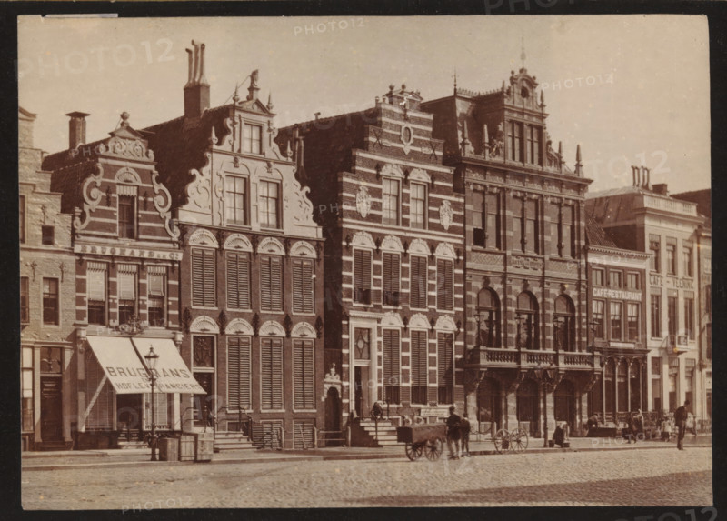 Buildings along the Grote Markt in Groningen, photograph, inscription: 'Groningen', photographer: Johannes Gerardus Kramer (possibly), Grote Markt (Groningen), c. 1880 - c. 1900, Groningen (city), paper, pasteboard, albumen print, height 96 mm x width 139 mm, square, circus, façade (of house or building), A row of ornate multi-story buildings with decorative facades and stepped gables lines a cobblestone street, each building featuring tall windows with shutters, arched doorways, and sculptural ornamentation on the upper levels; an awning extends from one storefront above the sidewalk, and wooden barrels and crates sit near the entrance; a few horse-drawn carts and a couple of horses stand on the street in front of the buildings, and small groups of people gather along the sidewalk and near doorways.