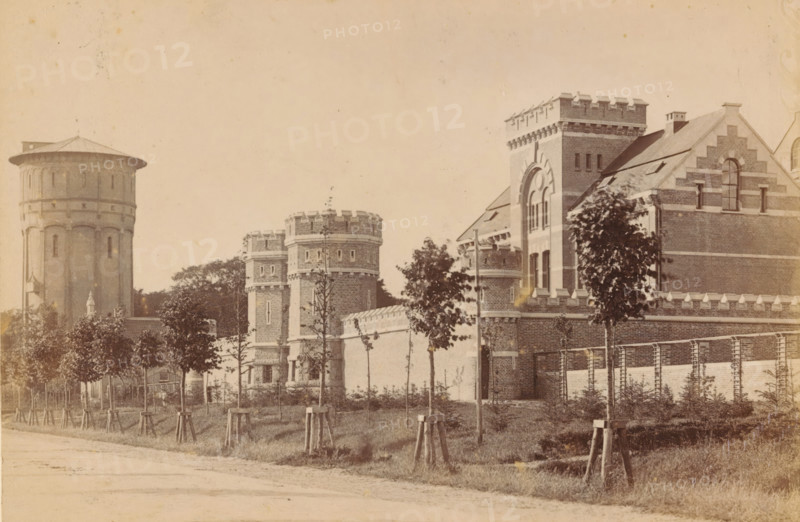 Exterior of the Cellular Penal Prison in Groningen, photograph, inscription: 'Groningen?', name: 'JG Kramer Groningen', photographer: Johannes Gerardus Kramer, Groningen (city), c. 1880 - c. 1900, paper, pasteboard, albumen print, height 96 mm x width 142 mm, façade (of house or building), prison, jail, Cellulaire Strafgevangenis, A sepia-toned street view shows a row of young trees with wooden supports along a grassy verge in front of a long brick wall topped with decorative stone battlements, behind which stand several brick buildings with castle-like architectural details including round towers with crenellated tops, a taller square tower with arched window openings, stepped gable ends, narrow vertical windows, and a large cylindrical water tower with a conical roof visible toward the left.