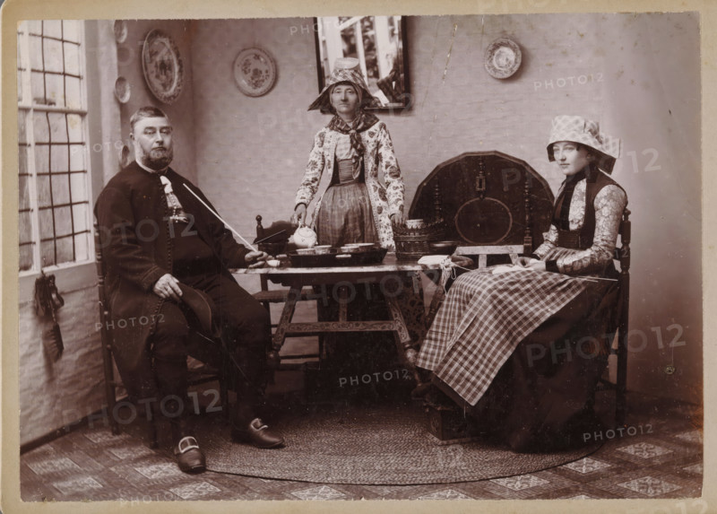 Portrait of an unknown man and two unknown women in traditional costumes from Hindeloopen, photograph, inscription: 'Th. M. Staas court photographer Leeuwarden Willemskade WL', inscription: 'Photograaf HJ de Koe Willemskade, W 1 Leeuwarden', photographer: Thomas Martin Staas, Hindeloopen, c. 1880 - c. 1900, paper, pasteboard, carbon print, height 99 mm x width 140 mm, family group, especially parents with their child(ren), folk costume, regional costume, interior, representation of a building, An indoor scene with three seated people around a low wooden table; a man in dark clothing with a beard sits on the left holding a hat and a long thin object, a woman in patterned traditional dress and a tall bonnet stands behind the table holding a teapot, and a young woman in patterned sleeves and a checked apron and bonnet sits on the right holding a book or paper on her lap; the table holds several bowls and a tray, and a basket and round wooden board lean against the wall behind them; the ro...