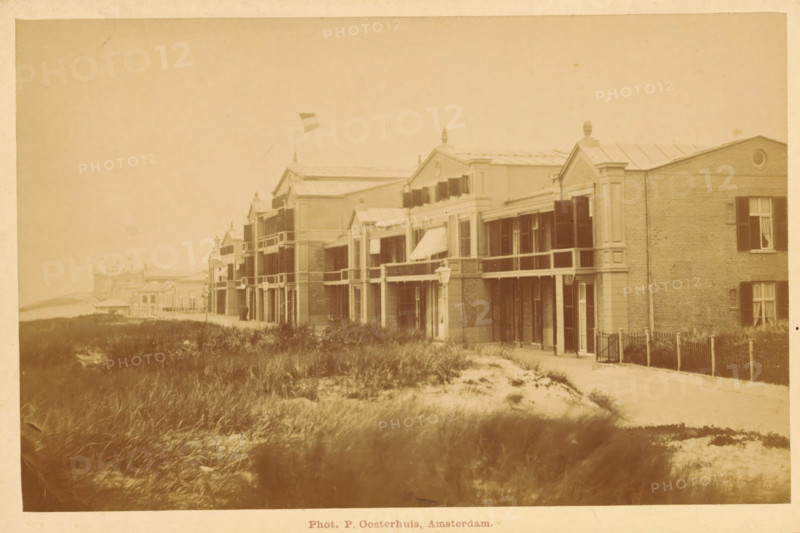 Facade of a building along the beach of Scheveningen, photograph, inscription: 'Scheveningen', photographer: Pieter Oosterhuis, Scheveningen, c. 1880 - c. 1900, paper, pasteboard, albumen print, height 94 mm x width 147 mm, beach, façade (of house or building), A sepia-toned photograph of a row of two-story buildings with balconies and shuttered windows facing a sandy, grassy foreground; some buildings have decorative pediments and finials on their roofs and one building displays a flag on a pole; a narrow path runs parallel to the buildings and a simple fence appears near one building; the foreground shows clumps of beach grass and patches of sand, and a distant shoreline is faintly visible.
