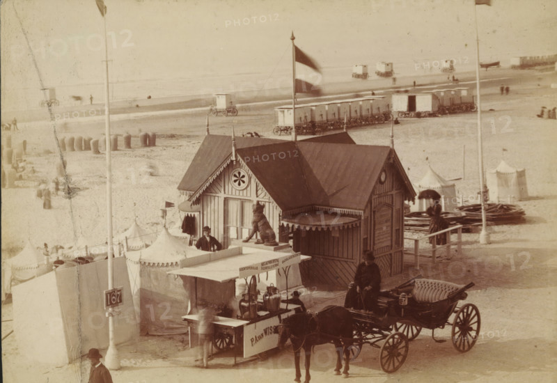 Wooden pavilion on the beach at Scheveningen, photograph, inscription: 'CP Wollrabe sen., The plate will be kept until reordered.', photographer: Carl Philip Wollrabe, Scheveningen, c. 1885 - c. 1900, paper, pasteboard, albumen print, height 100 mm x width 143 mm, beach (as place of recreation), four-wheeled, animal-drawn vehicle, eg: cab, carriage, coach, façade (of house or building), A sepia-toned beach scene shows a wooden pavilion with decorative trim and a peaked roof topped by a flag on a pole, a carved animal statue on a pedestal in front of the pavilion, and a man standing beside its entrance; in front of the pavilion is a cart with metal urns and a canopy, attached to a horse-drawn carriage with upholstered seating where a woman in dark dress and a hat sits, and another person stands nearby holding an umbrella; behind them are rows of small wheeled bathing huts on the sand and several tents with pointed tops, scattered people on the beach, piles of coiled material, and distan...