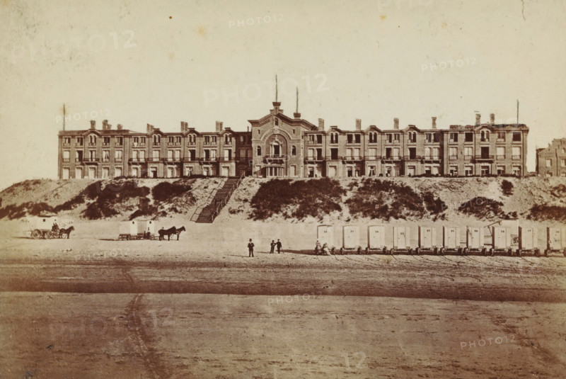 Exterior of the Oranje Hotel in Scheveningen, photograph, inscription: 'CP Wollrabe sen., The plate is kept until reordered.', photographer: Carl Philip Wollrabe, Scheveningen, c. 1885 - c. 1900, paper, pasteboard, albumen print, height 98 mm x width 146 mm, beach (as place of recreation), hotel, hostelry, inn, façade (of house or building), Oranjehotel, A wide, multi-story building with ornate architectural details stretches across a sandy embankment, centered by a taller central section with an arched feature and a narrow double staircase leading down the slope, flanked by lower wings with repeated windows and small balconies; grassy and shrub-covered dunes sit between the building and the beach, where a row of small rectangular beach carts on wheels lines the shore, several horses hitched to simple carts stand nearby, and a few people in period clothing are scattered along the sand, some standing and some near the carts.