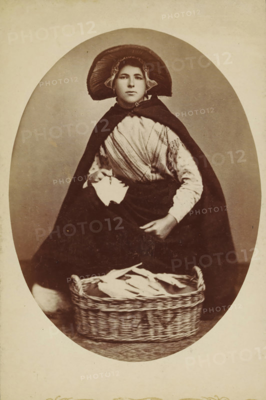 Portrait of an unknown woman in traditional costume, photograph, inscription: 'CP Wollrabe sen., The plate will be kept until reorder.', photographer: Carl Philip Wollrabe, South Holland, c. 1885 - c. 1900, paper, pasteboard, albumen print, height 149 mm x width 98 mm, adult woman, folk costume, regional costume, container made of plant material other than wood: basket, A sepia-toned portrait of a seated figure wearing a wide, rounded bonnet with ruffled edges and a dark cape draped over the shoulders, a patterned shawl visible across the chest and a long dark skirt covering the legs; one hand holds a folded piece of cloth while the other rests on the skirt, and a woven basket filled with folded linens sits on the floor in front of the figure against a plain oval background.