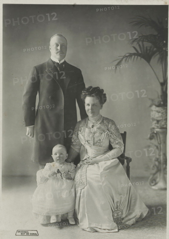 Portrait of Wilhelmina, Queen of the Netherlands, Hendrik of Mecklenburg-Schwerin and Juliana, photograph, Hendrik and Juliana. Bottom left 'ORIGINAL'., address: 'Guy de Coral & Co HOF-FOTOHANDEL AMSTERDAM-DEN HAAG.', photographer: Coral & Co., Guy de, Netherlands, 1909 - 1911, paper, pasteboard, photography, height 149 mm x width 106 mm, height 235 mm x width 146 mm, The title mentioned here refers to the highest title obtained by the person portrayed, not necessarily to the title the person portrayed bears in this image., Person, Hendrik (Prince of the Netherlands (1876-1934)), Juliana of the Netherlands, Wilhelmina of the Netherlands, A tall man in a dark formal coat stands behind a seated woman and a small child; the man has a moustache and wears a high-collared shirt with a tie. The woman sits on a wooden chair wearing a long, ornate dress with lace and embroidered details, a layered necklace, and her hair styled up; one hand rests on the child's shoulder and the other is in her l...