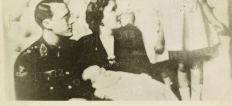 Portrait of Juliana, Queen of the Netherlands, Bernhard van Lippe-Biesterfeld, Beatrix, Irene, Princess of the Netherlands, and Margriet, photograph, Bernhard, Irene and Margriet in Ottawa., inscription: 'found in the field at Bocholtz L 1940-45 Signed H Dolmans 1980', Netherlands, 1943 - 1944, photographer, paper, photography, height 81 mm x width 113 mm, The title mentioned here refers to the highest title obtained by the person portrayed, not necessarily to the title the person portrayed bears in this representation., Person, Margriet (Princess of the Netherlands), Juliana of the Netherlands, North America, Ottawa, A grainy black-and-white photograph showing three seated figures and a bundled infant; on the left a person in a dark uniform with visible insignia on the shoulder and chest looks toward the center, wearing glasses and short hair; in the center a person with dark hair in a dark dress or coat faces slightly to the right, their face turned upward; on the right a lighter-ton...